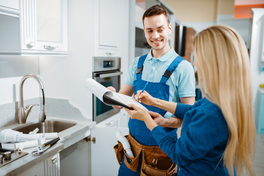 Plumber in uniform with satisfied customer in the kitchen signing receipt for completed work. Handyman with toolbag and tools.