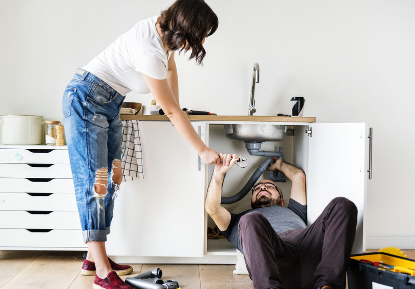 Couple fixing kitchen sink, man under the kitchen counter, woman handing him tools