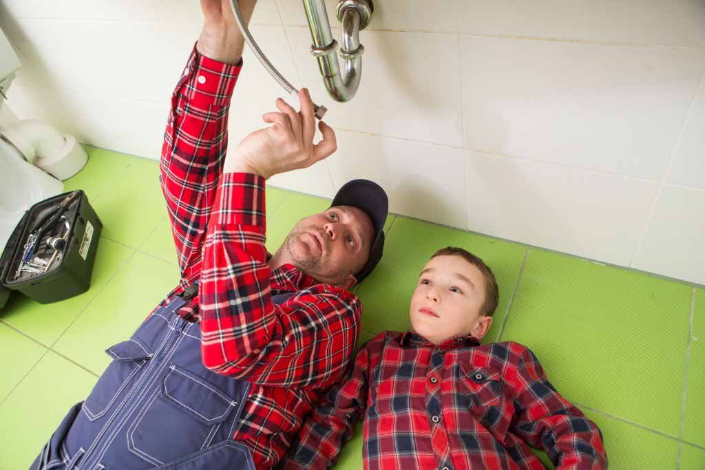 Man showing small boy the plumbing pipes underneath the sink area.