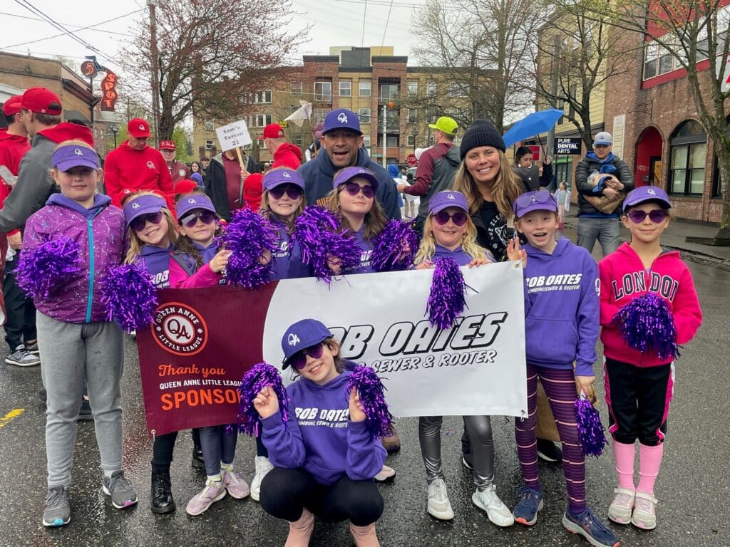 Queen Anne Little League youth team in purple uniforms posing with Bob Oates sponsorship banner at a local parade.
