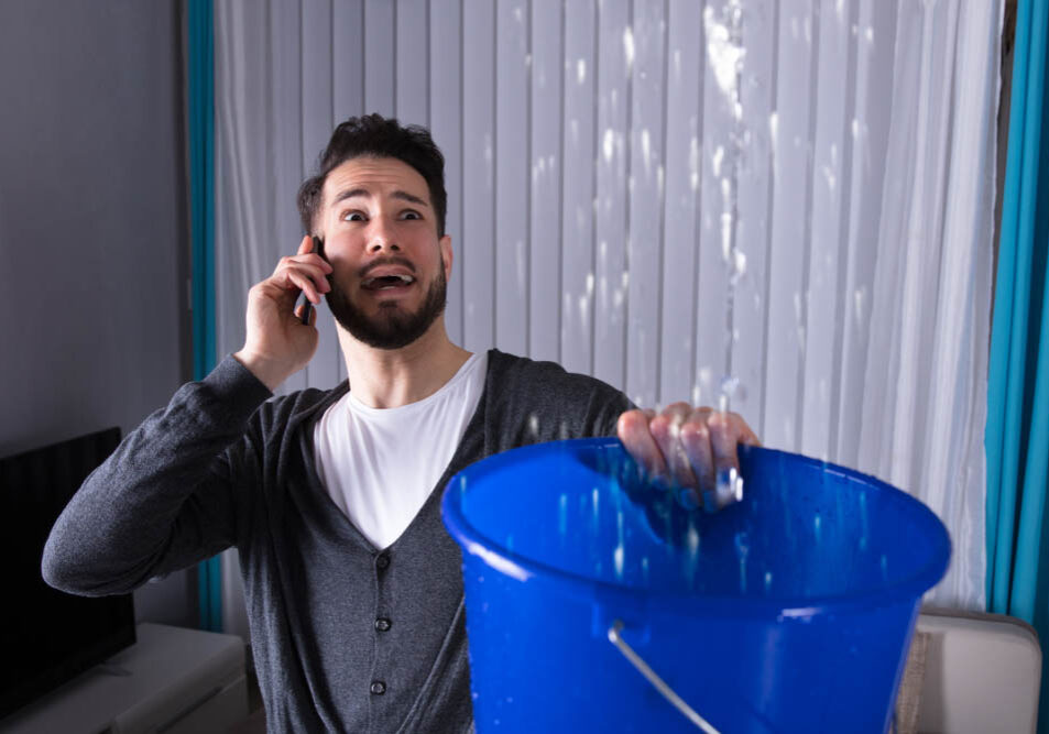 Worried young man calling plumber while leaking water is falling into bucket at home